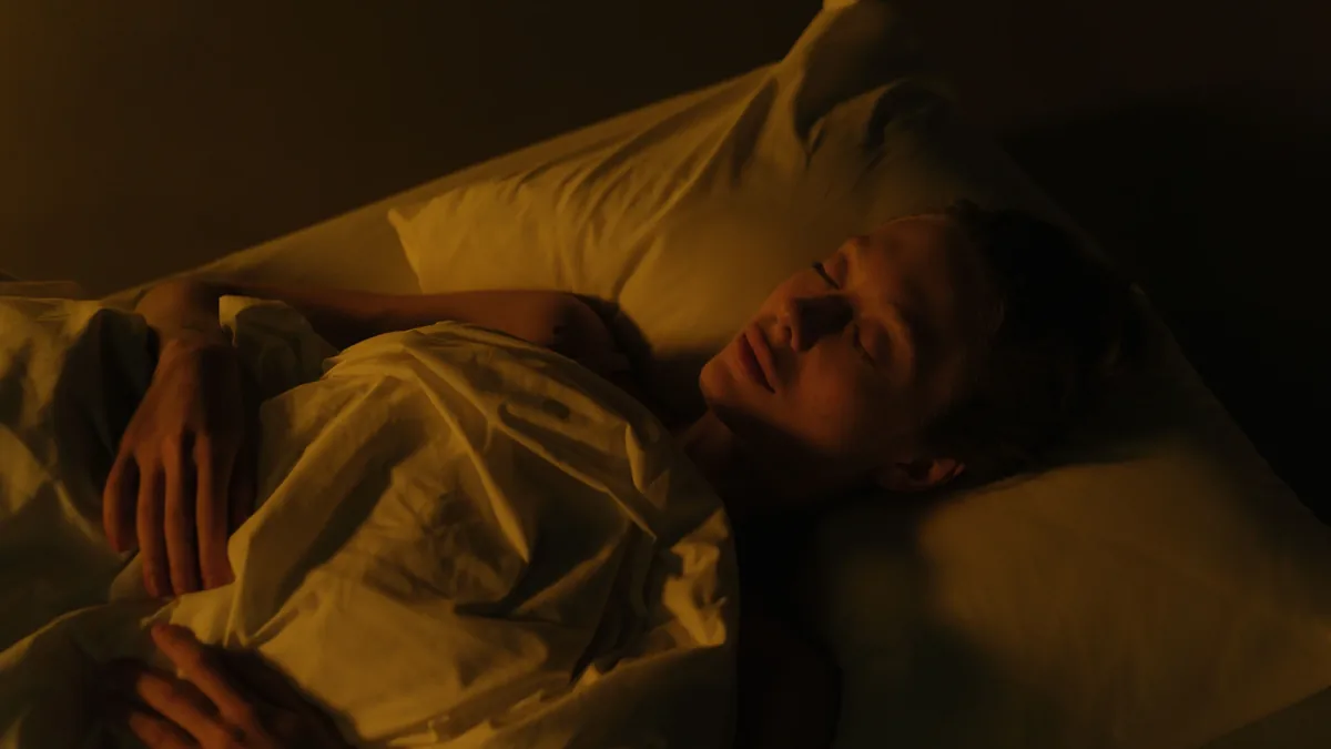 A person lying on their back in bed, covered by a sheet and resting their head on a pillow.