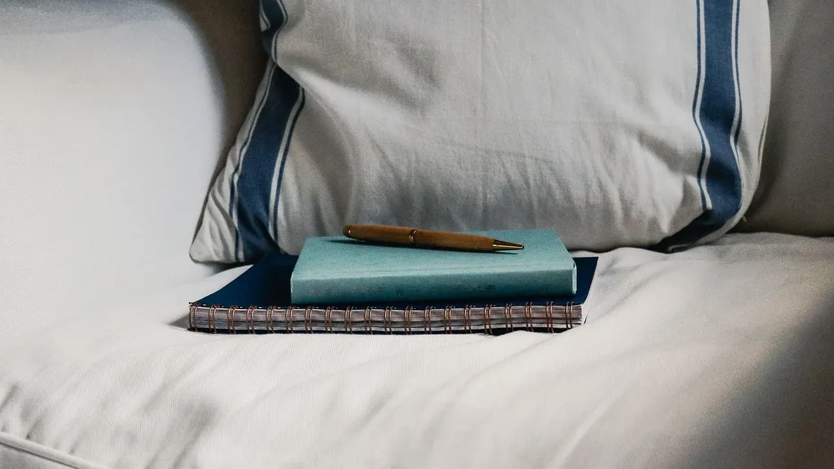 A stack of two books and a pen on a bed with white sheets and a blue and white pillow.