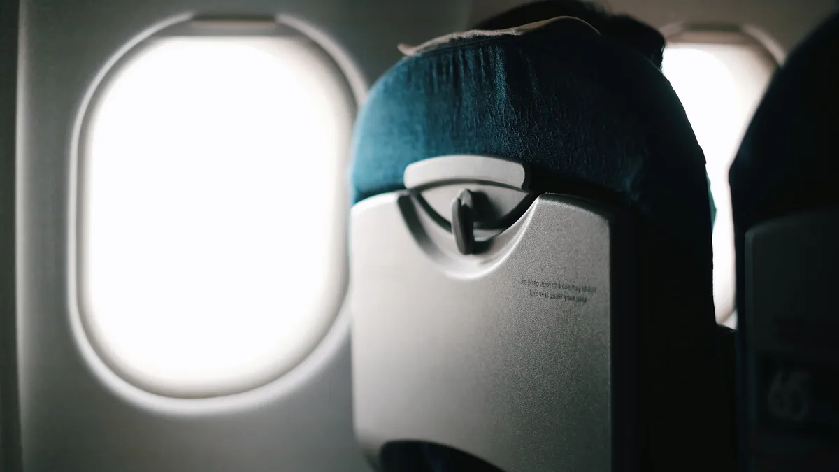 A person wearing a sleep mask on an airplane, with the window and seat visible in the background.