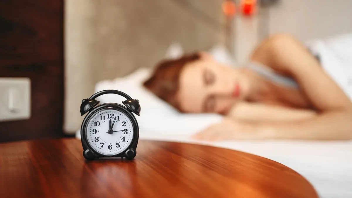 A woman lying on a bed, with an alarm clock on a bedside table in the foreground.