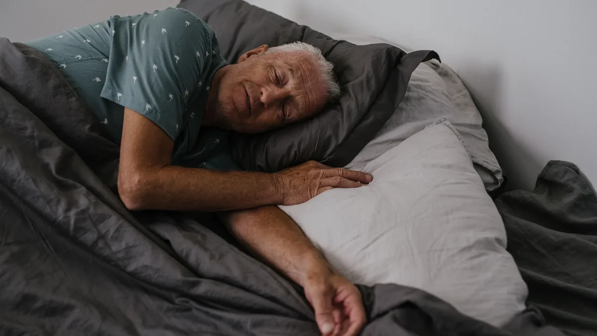 An elderly man is sleeping on a bed, lying on his side with his head resting on a pillow and his left arm bent at the elbow.