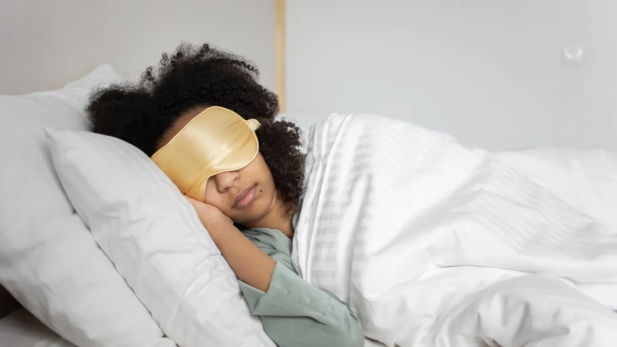 A woman is sleeping in bed, covered by a white blanket and wearing a beige sleep mask.