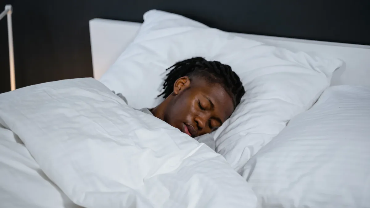 A person sleeping in bed, covered by a white duvet and surrounded by white pillows.
