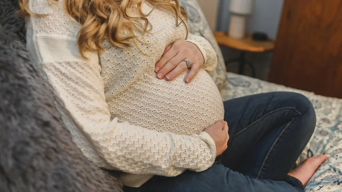 A pregnant woman sitting on a bed, with her hands on her stomach.