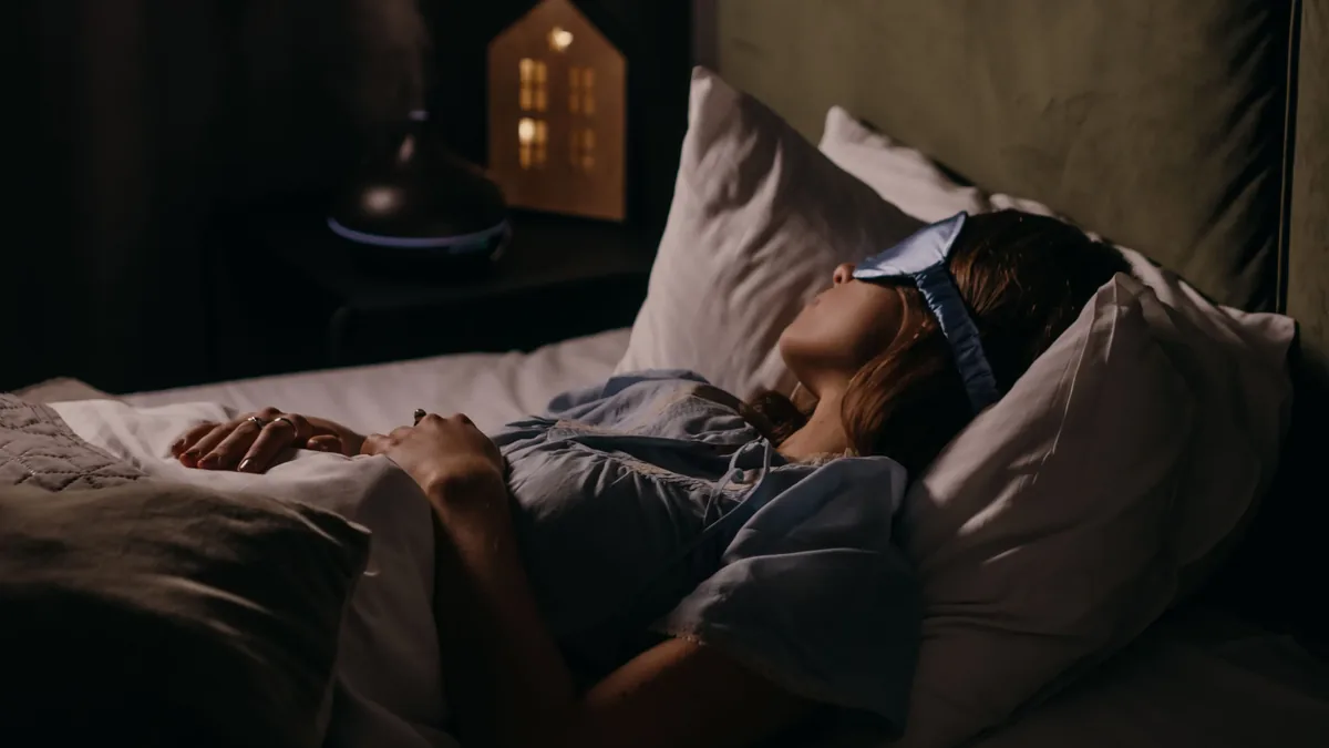 A woman is reclining on her back in bed, wearing a blue sleep mask and a blue shirt, with a white pillow supporting her head and a white she