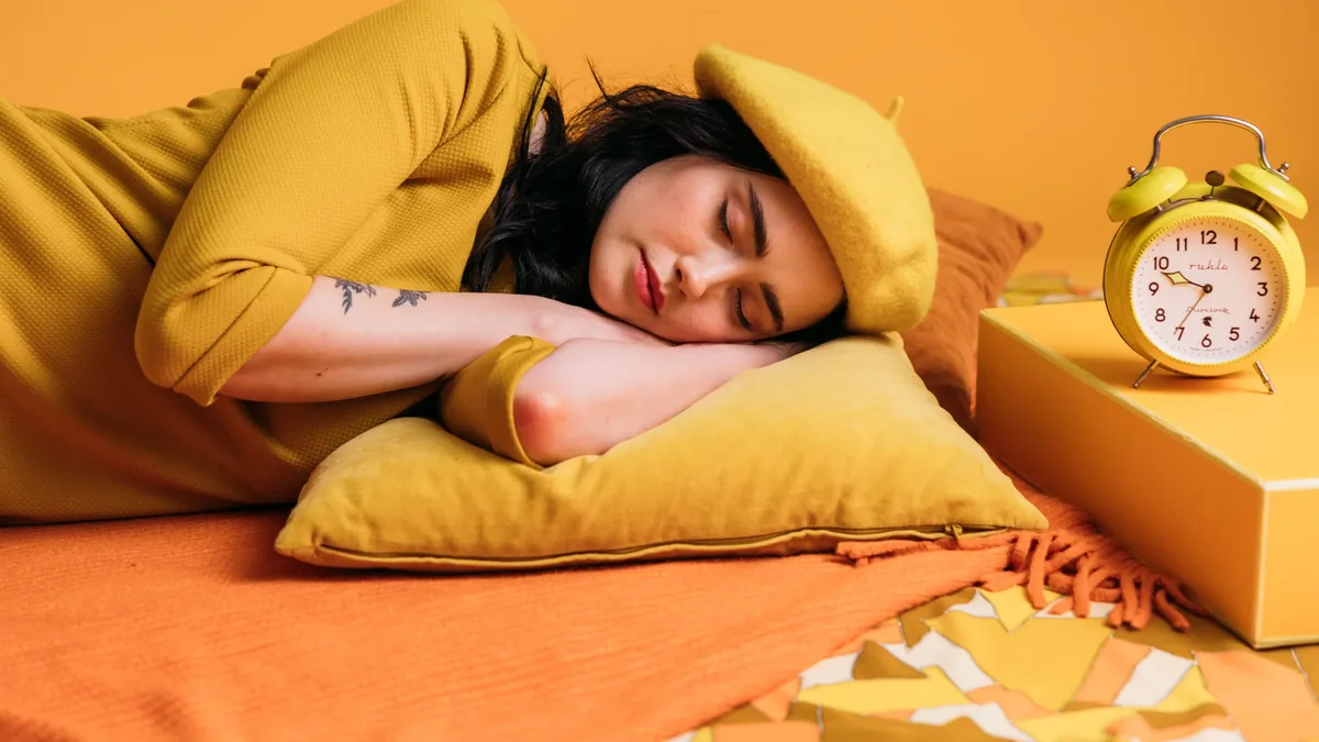 A woman sleeping on a bed with a yellow alarm clock and a yellow pillow.